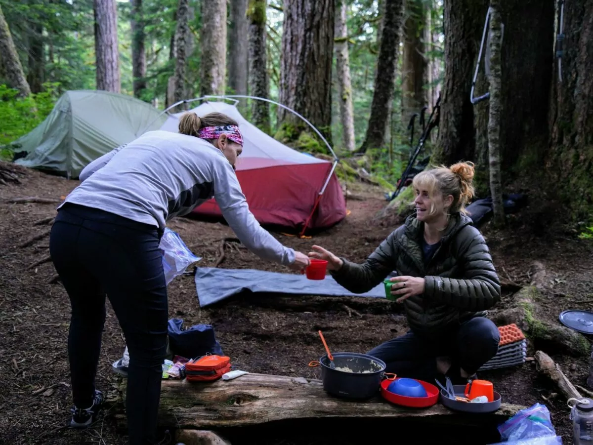 A guide hands a coffee cup to a guest at a wilderness campsite in the woods