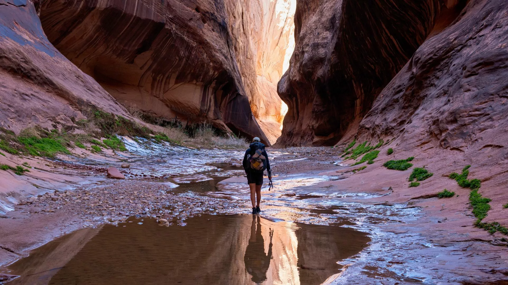 The author backpacks through shallow water in a Utah slot canyon, casting a reflection behind him