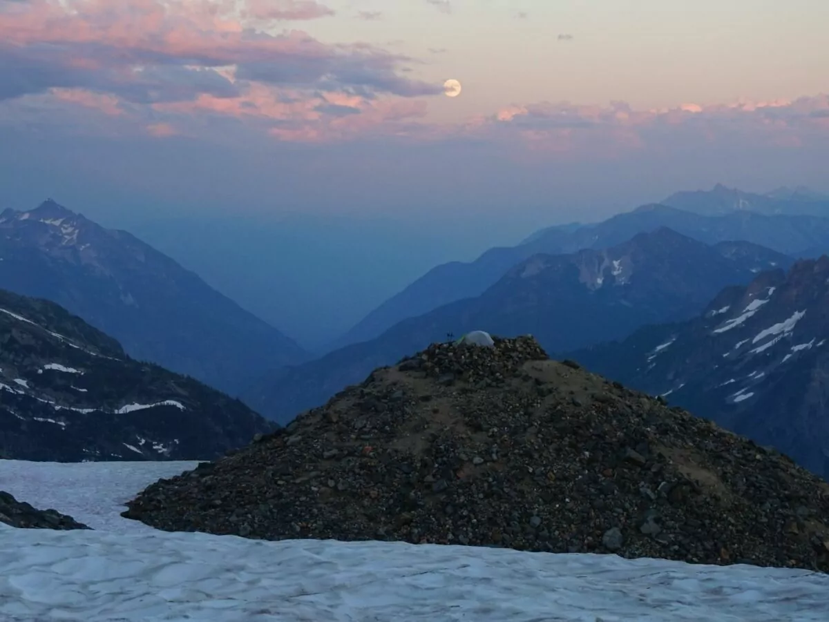 A tent sits on a rocky mountain top under a full moon and setting sun