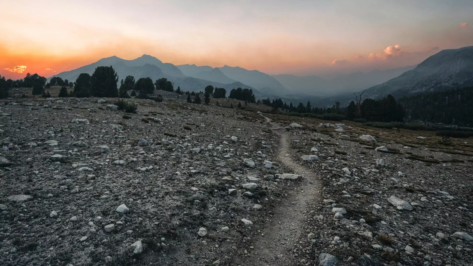 as the sun sets, a well worn hiking trail stretches across a rocky slope toward hazy mountains in the distance