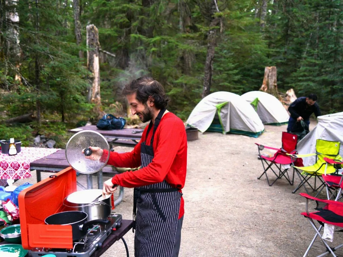 A man cooks in a campsite with tents and other camping equipment around him