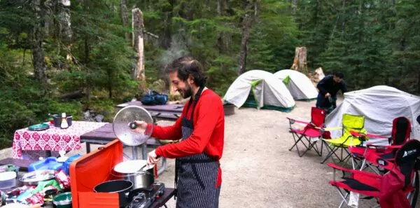 A man cooks in a campsite with tents and other camping equipment around him