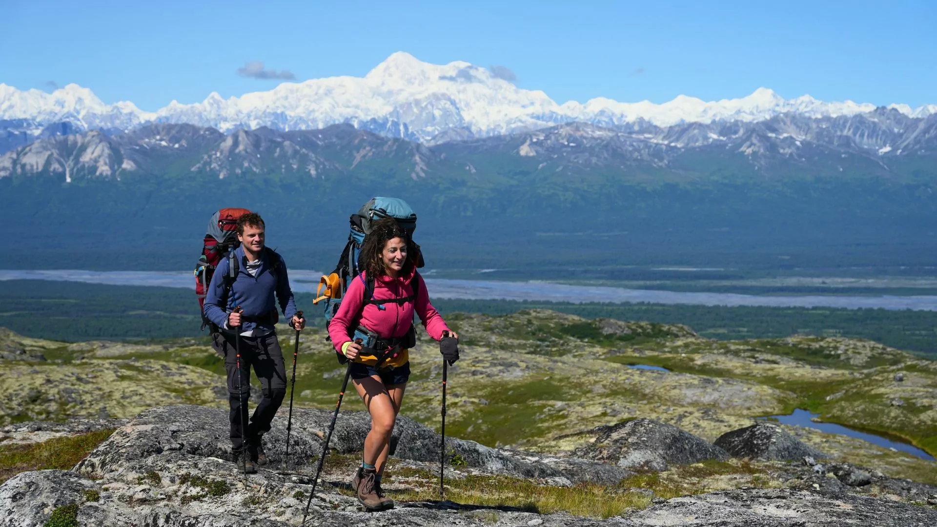 Two hikers with large deuter backpacks trek in front of a snowy mountain