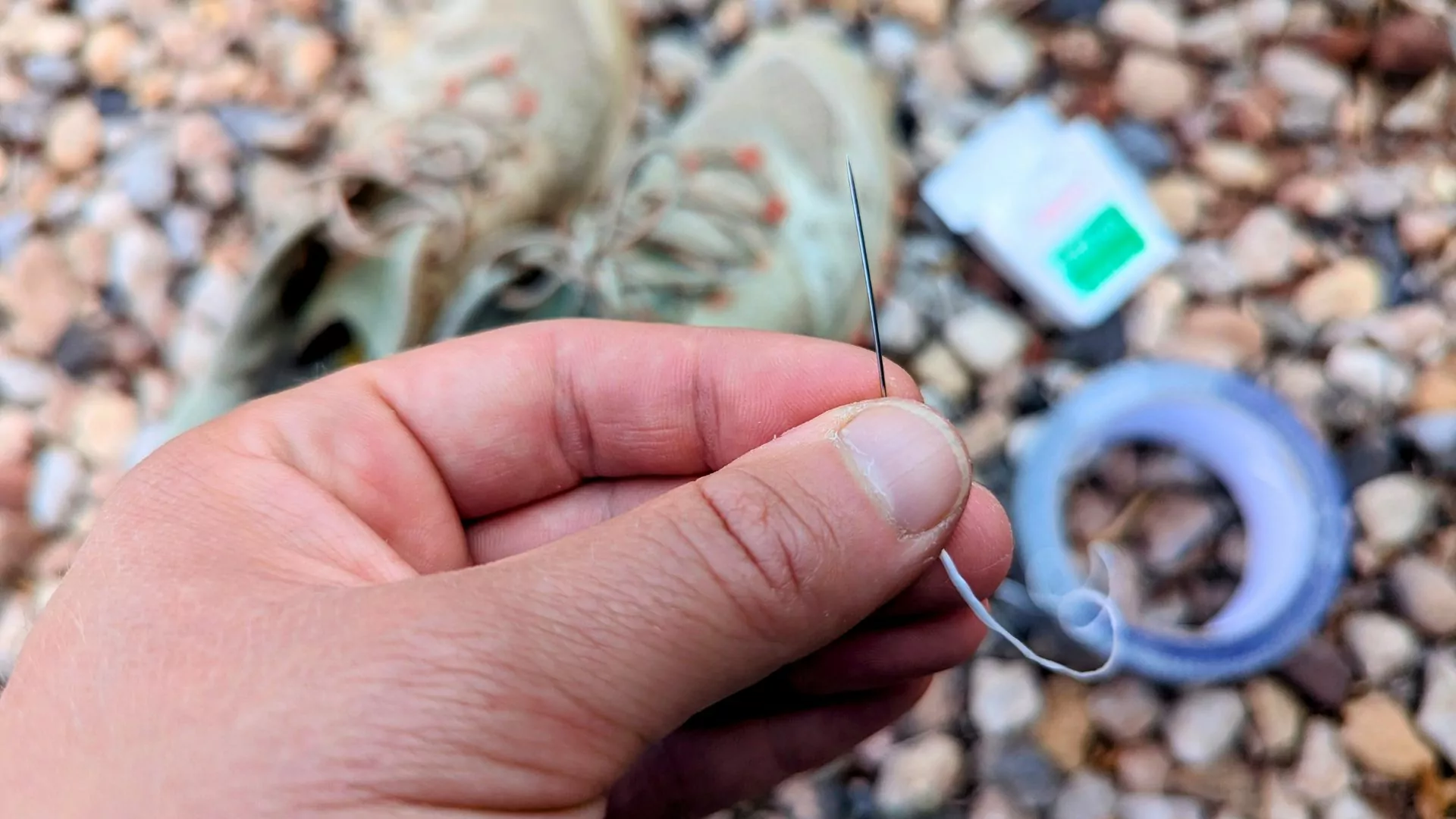 A needle threaded with dental floss is held next to a pair of shoes and a roll of duct tape
