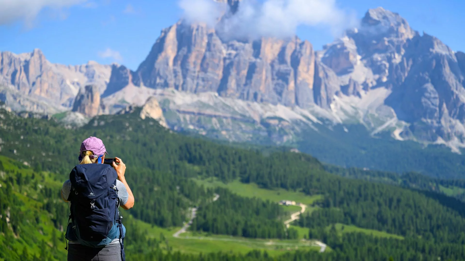 A hiker in the Dolomites wears a backpack and holds a camera while looking at the mountain view