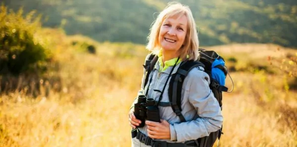 A smiling woman with binoculars and a daypack poses outside